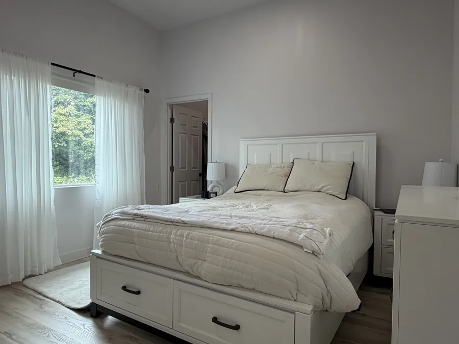 A bedroom with light curtains and a window onto green trees